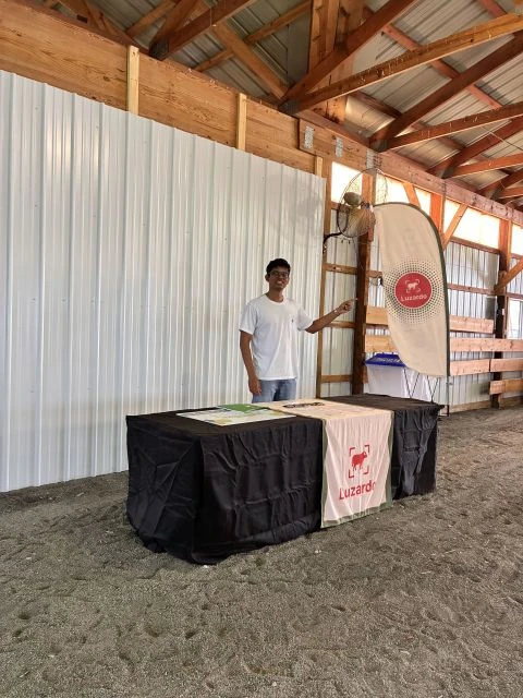 A member of the Luzardo team standing beside the Luzardo banner and booth table inside the exhibition barn